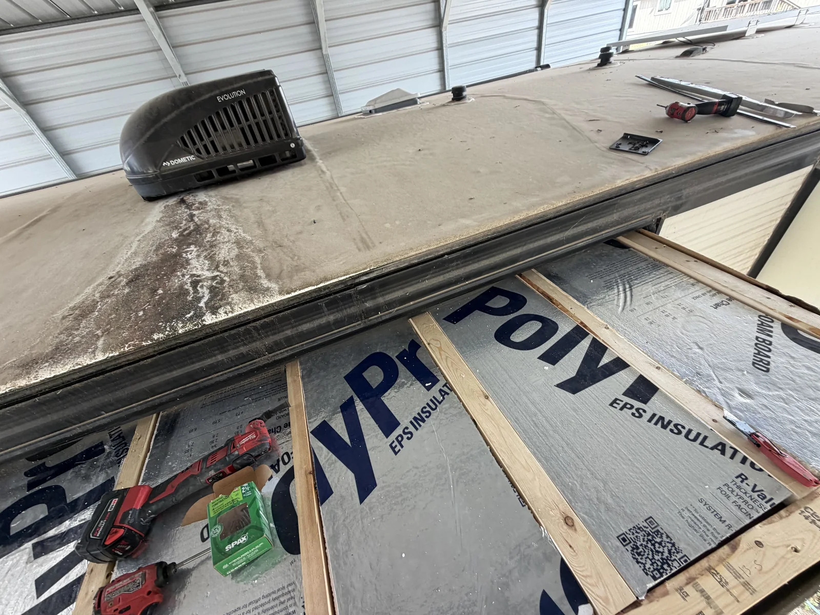 New insulation board laid on the roof framing of a travel trailer under a carport, with the rooftop AC unit in place, RV roof repair