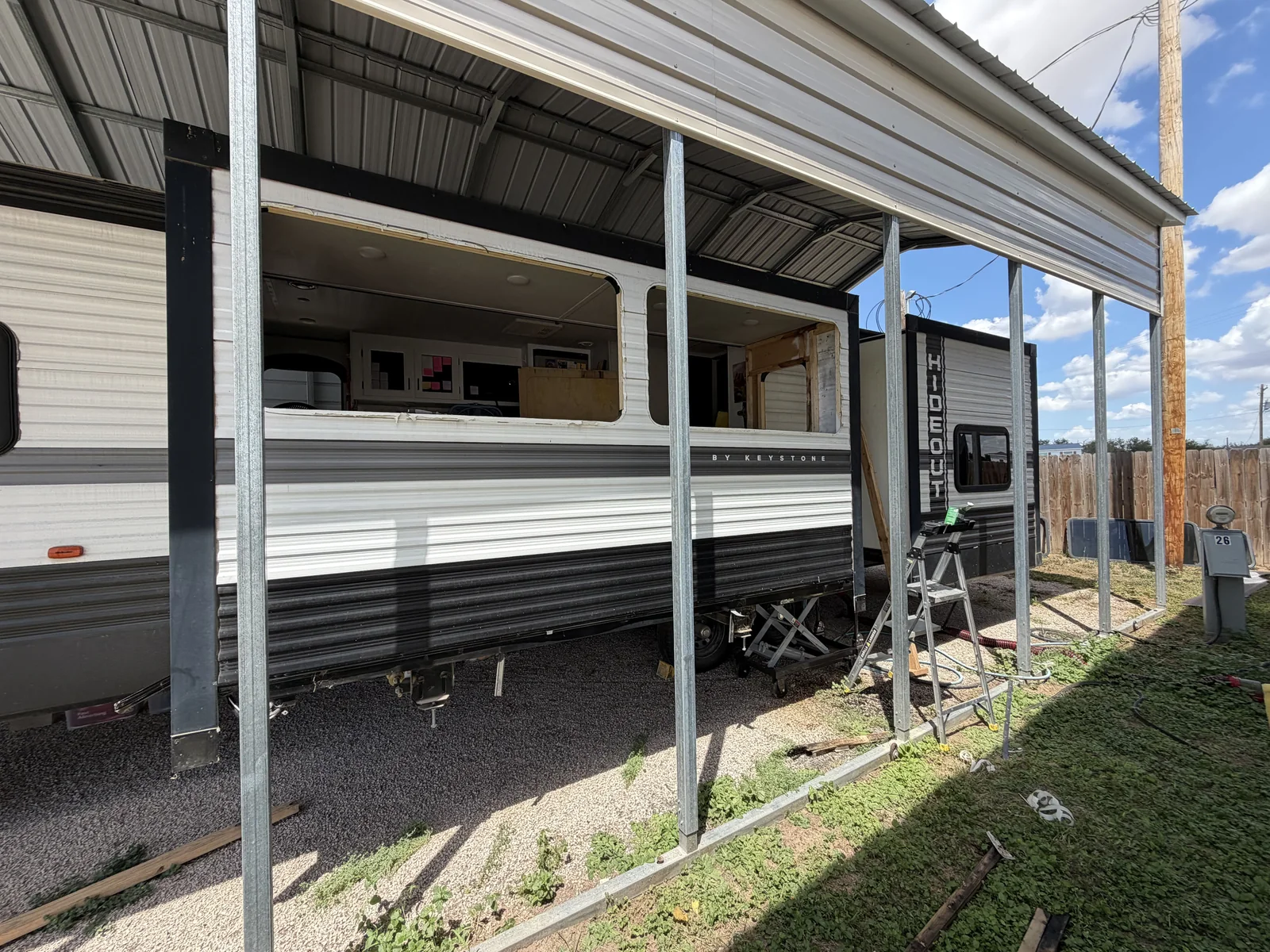 Travel trailer under a carport with a side wall opened up for a rebuild, a ladder set against it, RV body repair